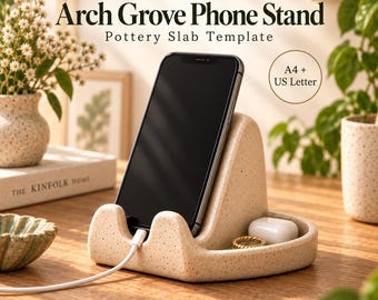 May include: A speckled beige ceramic phone stand with an arched back, holding a black smartphone connected to a white charging cable. The base features a small tray area containing a white earbud case and a gold ring. Text reads Arch Grove Phone Stand.