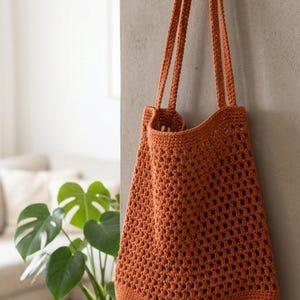 Peut inclure: Un sac fourre-tout en crochet orange brûlé présentant un motif ajouré à carrés sur le corps principal, une base solide et deux longues bretelles. Le sac est fabriqué en fil avec un point texturé visible sur toute la pièce.