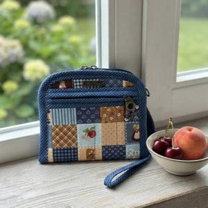 Puede incluir: Un bolso de mano de tela acolchada con un diseño de patchwork de cuadrados azules, marrones y crema con varios patrones, incluyendo cerezas y cuadros vichy. El bolso tiene un cierre de cremallera, un tirador de metal y una correa de muñeca.