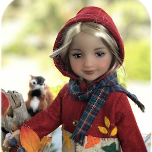 May include: A doll wearing a red hooded jacket with a plaid scarf and a skirt. The jacket has a patch with embroidered leaves and a button closure. The doll has blonde hair and brown eyes.