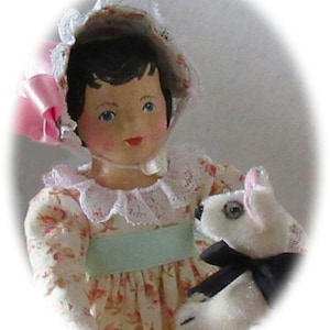 May include: A porcelain doll wearing a pink and white bonnet and a floral dress holds a white stuffed animal with black bow tie.