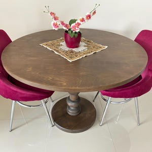Peut inclure: Une table à manger ronde en bois avec une finition brun foncé et un pied central, accompagnée de deux chaises en velours magenta aux pieds en métal argenté. Un napperon carré à motifs est posé sous un pot magenta avec des orchidées blanches et roses.