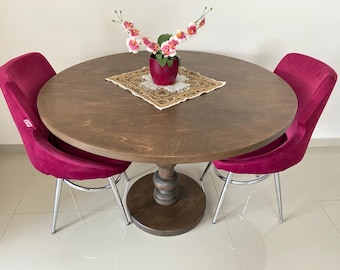 Pode incluir: Uma mesa de jantar redonda de madeira com acabamento castanho escuro e base central de pedestal, acompanhada por duas cadeiras de veludo magenta com pernas de metal prateado. Um naperon quadrado com padrões está sob um vaso magenta com orquídeas brancas e rosas.