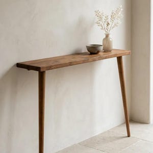 Pode incluir: Uma mesa consola estreita de madeira com um tampo retangular e quatro pernas c&oacute;nicas. Na superf&iacute;cie encontram-se uma pequena ta&ccedil;a de cer&acirc;mica cinzenta e um vaso de vidro transparente com caules bot&acirc;nicos brancos secos.