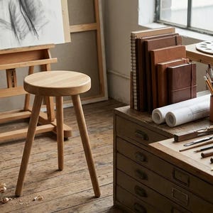 May include: A light-colored wooden stool with three round legs and a circular seat, positioned in an art studio setting featuring a wooden easel, a set of brown leather-bound journals, drawing tools, and a wooden cabinet with drawers.