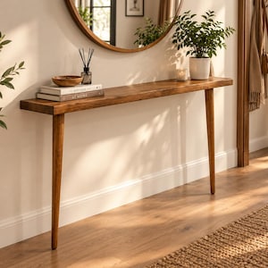 Pode incluir: Uma mesa consola de madeira longa e estreita com um acabamento liso castanho m&eacute;dio e quatro pernas c&oacute;nicas. A superf&iacute;cie da mesa cont&eacute;m dois livros empilhados intitulados Kinfolk e At Home, uma ta&ccedil;a de madeira, um difusor e uma planta.