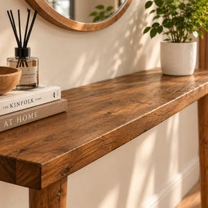 Pode incluir: Uma mesa consola retangular de madeira comprida com acabamento de gr&atilde;o natural e pernas c&oacute;nicas. Na superf&iacute;cie encontram-se dois livros empilhados intitulados The Kinfolk Home e At Home, uma ta&ccedil;a de madeira com chaves e um difusor de varetas.