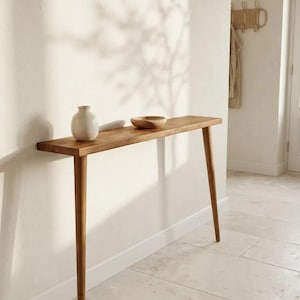 May include: A narrow wooden console table with four tapered legs, featuring a smooth rectangular top. On the surface sits a small, off-white ceramic vase and a shallow, round wooden bowl. The table is constructed from light brown wood.