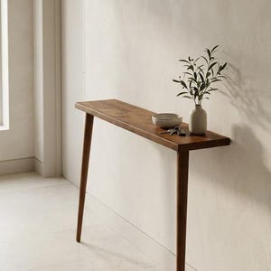 Pode incluir: Uma mesa consola de madeira estreita e retangular com quatro pernas c&oacute;nicas. Na superf&iacute;cie encontram-se uma pequena ta&ccedil;a de cer&acirc;mica clara, um conjunto de chaves de metal e um vaso de cer&acirc;mica clara com um ramo de planta verde.