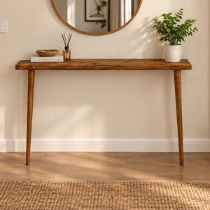 Pode incluir: Uma mesa consola retangular de madeira com acabamento castanho natural e quatro pernas c&oacute;nicas. O tampo da mesa cont&eacute;m um livro branco, uma pequena ta&ccedil;a de madeira, um difusor de varetas de vidro e uma planta verde num vaso.