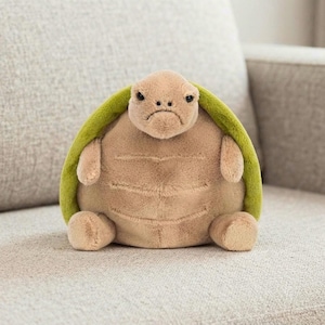 May include: A plush turtle toy featuring a soft, tan-colored body with a rounded, textured shell in a vibrant lime green. The stuffed animal has a small head with dark, bead-like eyes and a neutral expression, with four short, stubby limbs.