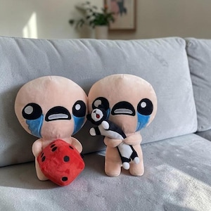 May include: Two light peach-colored plush figures with large black eyes and blue tear-shaped patches on their cheeks. One figure holds a red plush die with black dots, while the other holds a small black and white plush cat-like creature.
