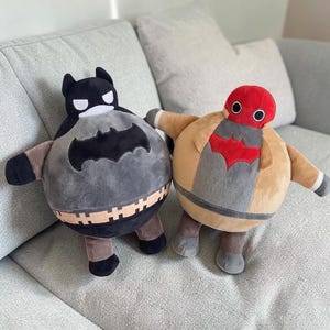 Peut inclure: Deux peluches rondes aux designs de personnages. Le jouet de gauche est noir et gris avec un emblème de chauve-souris et un masque. Le jouet de droite est beige et rouge avec un emblème rouge et une tête rouge. Corps sphériques et textures douces.