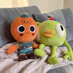 Puede incluir: Dos peluches: un personaje naranja de cabeza redonda, grandes ojos negros, un pequeño sombrero azul y traje de explorador, y una criatura verde con un gran ojo blanco, un pico amarillo y una pequeña hélice colorida en la cabeza.