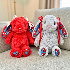 Könnte beinhalten: Zwei Plüschhasen, einer leuchtend rot und einer hellgrau, mit langen Schlappohren, die mit blauem Stoff mit Spider-Man-Motiven gefüttert sind. Der rote Hase hat weiße Bänder, während der graue Hase rote Bänder an den Ohren trägt.