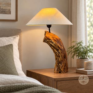 May include: A table lamp featuring a natural, curved wooden log base with a textured, golden-brown finish. The lamp is topped with a wide, off-white, conical fabric lampshade that emits a warm, soft glow from the light bulb underneath.