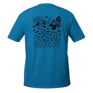 Puede incluir: Una camiseta azul brillante de manga corta con un estampado gráfico negro en la espalda. El diseño muestra una gran bandada de pájaros, una rama de árbol y una figura de personaje, con el texto FLOCK VS FLOCK en un borde rectangular.