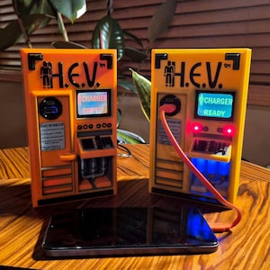 May include: Two orange and yellow H.E.V. charging stations with black text and icons. One unit displays CHARGER EMPTY, the other CHARGER READY with red and blue lights. An orange charging cable connects the ready unit to a black smartphone.