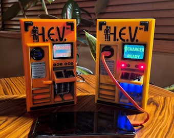 Pode incluir: Duas estações de carregamento H.E.V. laranja e amarelas com texto e ícones pretos. Uma unidade exibe CHARGER EMPTY, a outra CHARGER READY com luzes vermelhas e azuis. Um cabo de carregamento laranja conecta a unidade pronta a um smartphone preto.