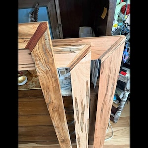 May include: A wooden picture frame featuring light tan wood with dark brown grain patterns and streaks. The corners of the frame are accented with contrasting dark reddish-brown wood inserts, creating a distinct geometric mitered joint design.