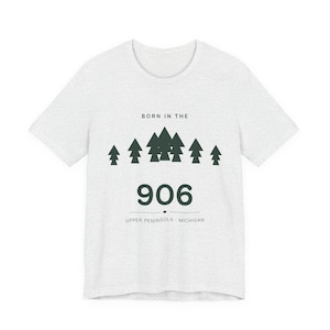 Può includere: Una t-shirt a maniche corte grigio melange chiaro con una grafica verde scuro di alberi di pino sopra il testo BORN IN THE 906 e UPPER PENINSULA MICHIGAN. Il design include una linea orizzontale che separa i numeri dal testo della località.