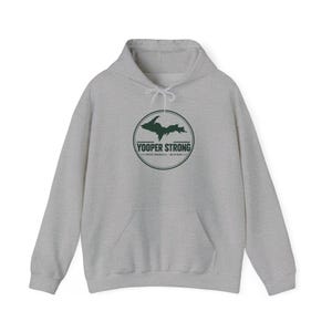 Può includere: Una felpa con cappuccio grigio chiaro caratterizzata da una grafica circolare verde scuro sul petto. La grafica raffigura la sagoma della penisola superiore del Michigan sopra il testo YOOPER STRONG e UPPER PENINSULA MICHIGAN in un cerchio.