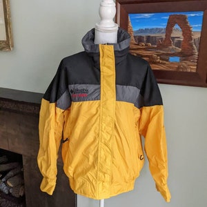 Könnte beinhalten: Eine gelbe, schwarze und graue Jacke von Columbia Sportswear mit Reißverschluss. Die Jacke hat ein Logo auf der linken Brust, auf dem "Columbia Sportswear" steht.