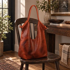 May include: A cognac-colored leather hobo bag with a slouchy silhouette, featuring two long shoulder straps that are knotted at the top attachment points. The bag has a smooth, polished finish and a deep, open main compartment.