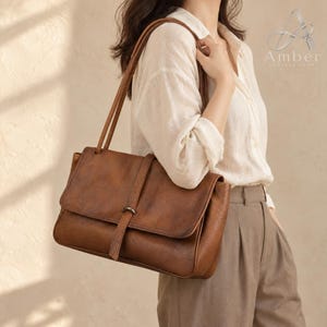 May include: A brown leather shoulder bag with a rectangular flap, a central vertical strap closure with a metal loop, and two thin shoulder straps. The bag features a textured surface and is branded with the text Amber Leather Shop.