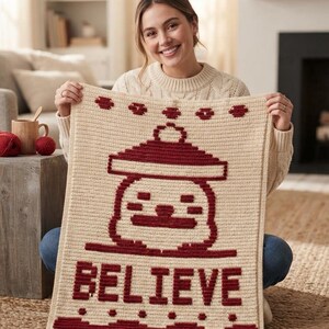May include: A rectangular crochet blanket in cream with a dark red graphic design. The pattern features a central ornament shape above the word BELIEVE in block letters, with decorative dotted borders at the top and bottom edges of the textile.