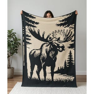 May include: A rectangular woven blanket featuring a high-contrast black and cream pixelated design of a large moose standing in a forest setting with pine trees and a textured, grid-like fabric construction throughout the entire piece.