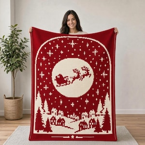 Puede incluir: Una manta de ganchillo rectangular con fondo rojo intenso y una escena invernal blanca. El diseño muestra la silueta de Santa Claus en un trineo tirado por renos volando frente a una gran luna blanca, sobre un pueblo nevado.