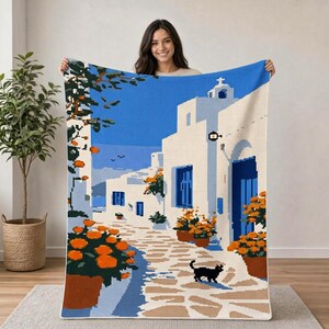 May include: A rectangular throw blanket featuring a pixelated Mediterranean scene with white buildings, blue doors, and windows. The design includes orange potted flowers, a stone pathway, and a small black cat, all set against a bright blue sky.