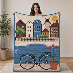 May include: A woven throw blanket featuring a pixelated cityscape with colorful houses, a windmill, and a bright yellow sun. The design includes a blue canal, a black bicycle, and a terracotta pot with three red tulips, all bordered by a blue trim.