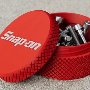 May include: A bright red, round, knurled storage container with the white Snap-on logo embossed on the lid. The lid is tilted open, revealing a collection of silver-coloured metal socket head cap screws stored inside the container.