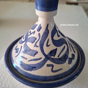 Peut inclure: Un tajine en céramique avec un couvercle conique orné de motifs calligraphiques bleus peints à la main sur un fond crème. Le couvercle possède une poignée cylindrique au sommet et le texte pottery Mosaic Art est visible sur le côté droit.