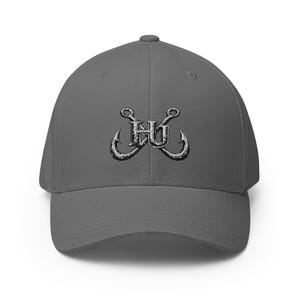Op de afbeelding: Een antracietgrijze baseballpet met een geborduurd ontwerp op de voorzijde. Het borduursel toont twee gekruiste, zilverkleurige vishaken achter de letters HU, uitgevoerd in een verweerd, zwart-wit lettertype met een versleten look.