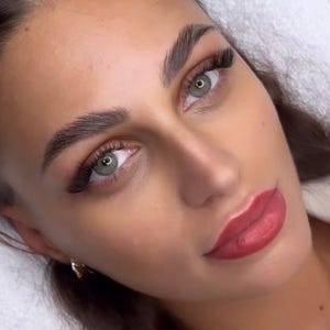 Könnte beinhalten: Ein Gesicht mit hellgrünen Augen, dunkel geformten Augenbrauen und langen schwarzen Wimpernverlängerungen. Die Lippen sind mit einem matten roten Pigment geschminkt und ein kleiner goldener Ohrring ist am Ohrläppchen sichtbar. Die Haut ist ebenmäßig.