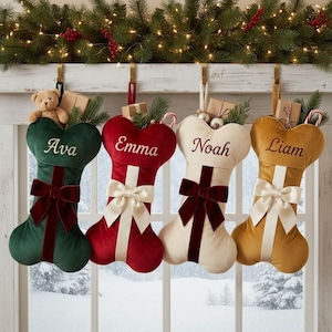 May include: Four bone-shaped velvet Christmas stockings in dark green, red, cream, and gold, each featuring a central ribbon with a bow and a personalized name embroidered in script. The stockings are filled with small gifts and greenery.