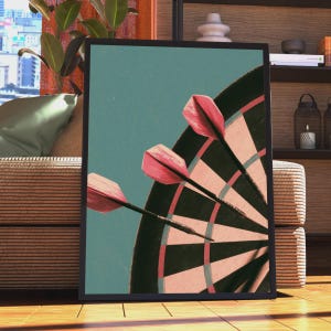 May include: A framed wall art print featuring a close-up view of three pink darts embedded in a dartboard. The dartboard has alternating black and light pink segments against a solid teal background. The frame is thin and black.
