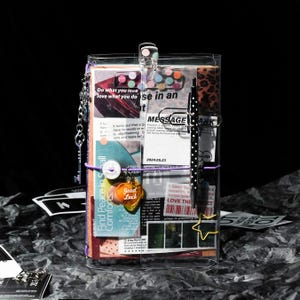 May include: A clear plastic journal cover containing a collage of printed papers, text, and images. It features a purple elastic band, a silver key charm, a black polka-dot pen, a yellow star paperclip, and a heart-shaped Good Luck charm.