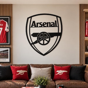 May include: A brown leather sofa with black and red throw pillows featuring the Arsenal cannon logo. A wooden coffee table holds books titled Arsenal, The Beautiful Game, and Football, alongside a black mug with the Arsenal crest logo.