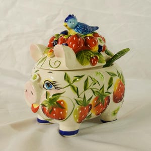 May include: A ceramic pig-shaped jar with a lid featuring a blue bird perched atop a cluster of red strawberries. The jar is cream-colored and decorated with hand-painted green leaves and red strawberries around the body and lid.