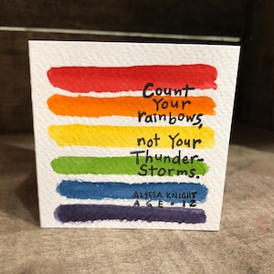 May include: A square white card featuring six horizontal watercolor stripes in red, orange, yellow, green, blue, and purple. Black handwritten text reads: Count Your rainbows, not Your Thunder-Storms. Below is: ALYSSA KNIGHT AGE 12.