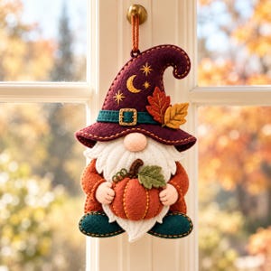 Op de afbeelding: Een vilt gnome ornament met een donkerpaarse puntmuts voorzien van geborduurde maan- en stermotieven, een turquoise hoedband met gesp en herfstbladeren. De gnome heeft een witte baard, oranje mouwen, turquoise schoenen en houdt een oranje pompoen vast.