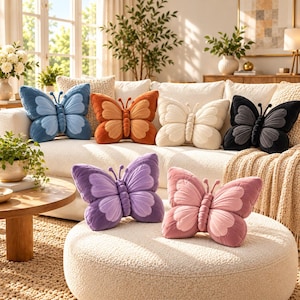 May include: A collection of six plush butterfly-shaped throw pillows in various colors including blue, orange, cream, black, purple, and pink. Each pillow features a central body, two antennae, and four distinct wing sections with soft textures.