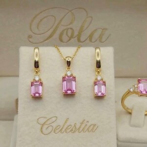 Könnte beinhalten: Ein Schmuckset bestehend aus einer Goldkette, zwei Ohrhängern und einem Ring, jeweils besetzt mit einem rechteckigen rosa Edelstein und kleinen klaren Akzentsteinen. Die Stücke liegen in einer cremefarbenen Schachtel mit dem Aufdruck Pola Celestia.