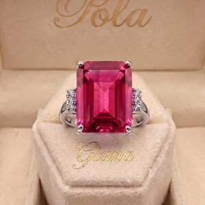 Könnte beinhalten: Ein silberfarbener Ring mit einem großen, smaragdgeschliffenen, leuchtend pinkfarbenen Edelstein als Herzstück. Die Ringschiene ist auf beiden Seiten mit kleinen, klaren, rund geschliffenen Steinen besetzt. Der Ring liegt in einer beigen Box mit dem Text Pola und Giana.