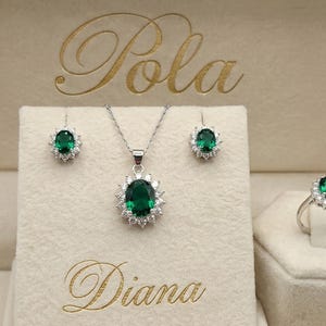 May include: A jewelry set featuring a necklace, two earrings, and a ring, each with an oval-cut emerald green stone surrounded by small clear stones. The set is displayed in a beige box with the text Pola Diana printed in gold script lettering.