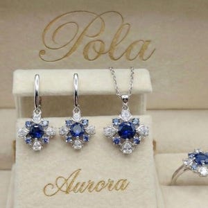 May include: A jewellery set featuring a pair of drop earrings, a pendant necklace, and a ring, all crafted with a central round blue stone surrounded by smaller blue and clear stones in a starburst pattern, presented in a beige box labelled Pola Aurora.